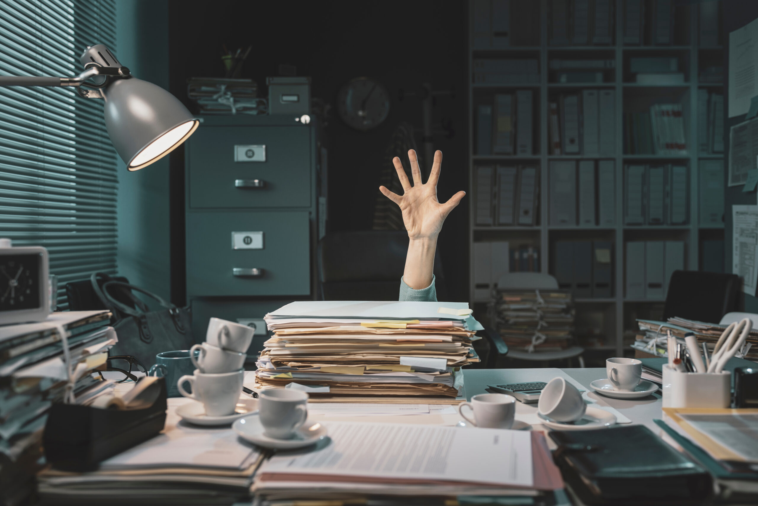 Overwhelmed Office Worker – A hand reaches out from behind a huge stack of paperwork in a cluttered office.
