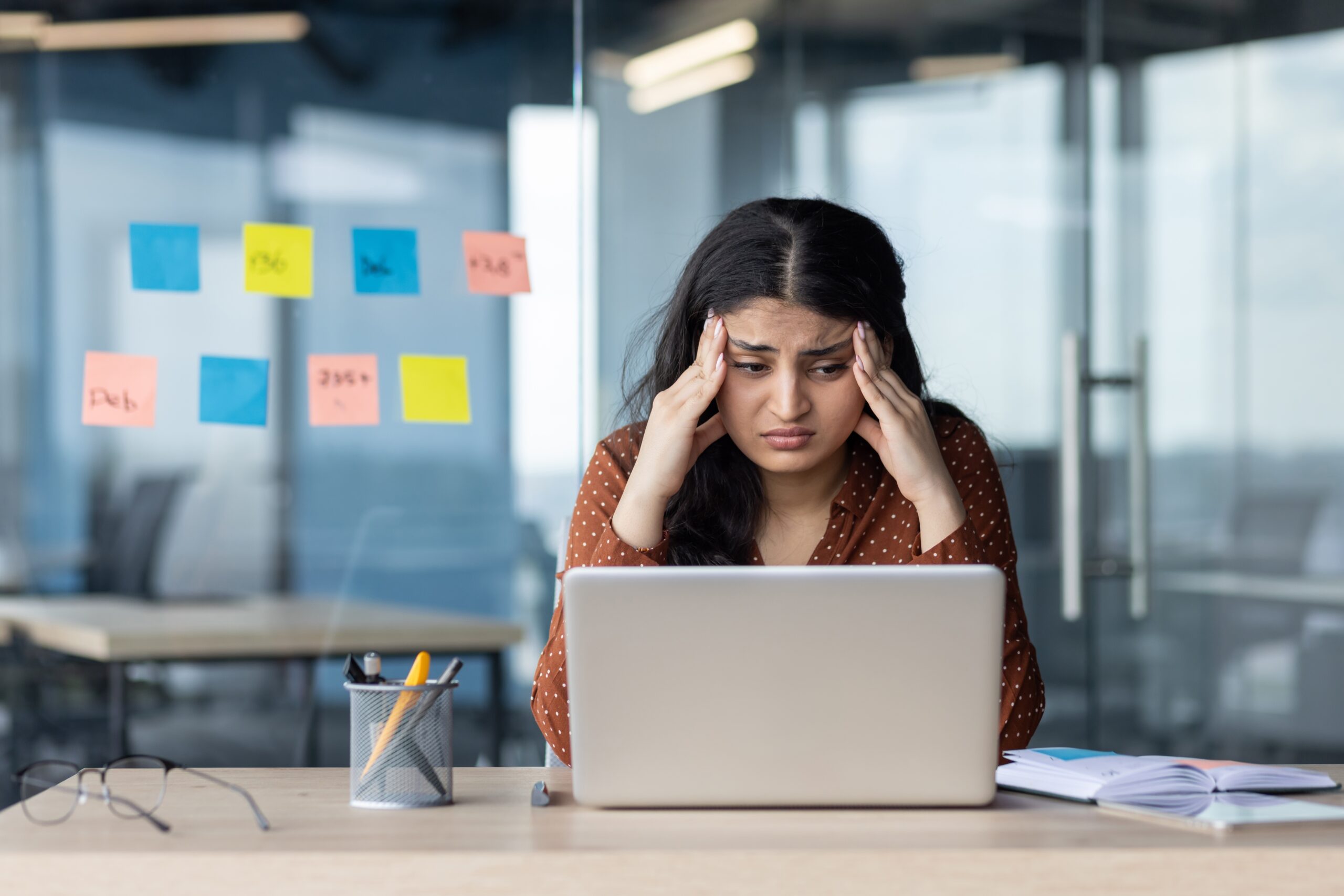 Stressed Woman at Work – A woman looks stressed at her laptop, surrounded by sticky notes in a modern office.