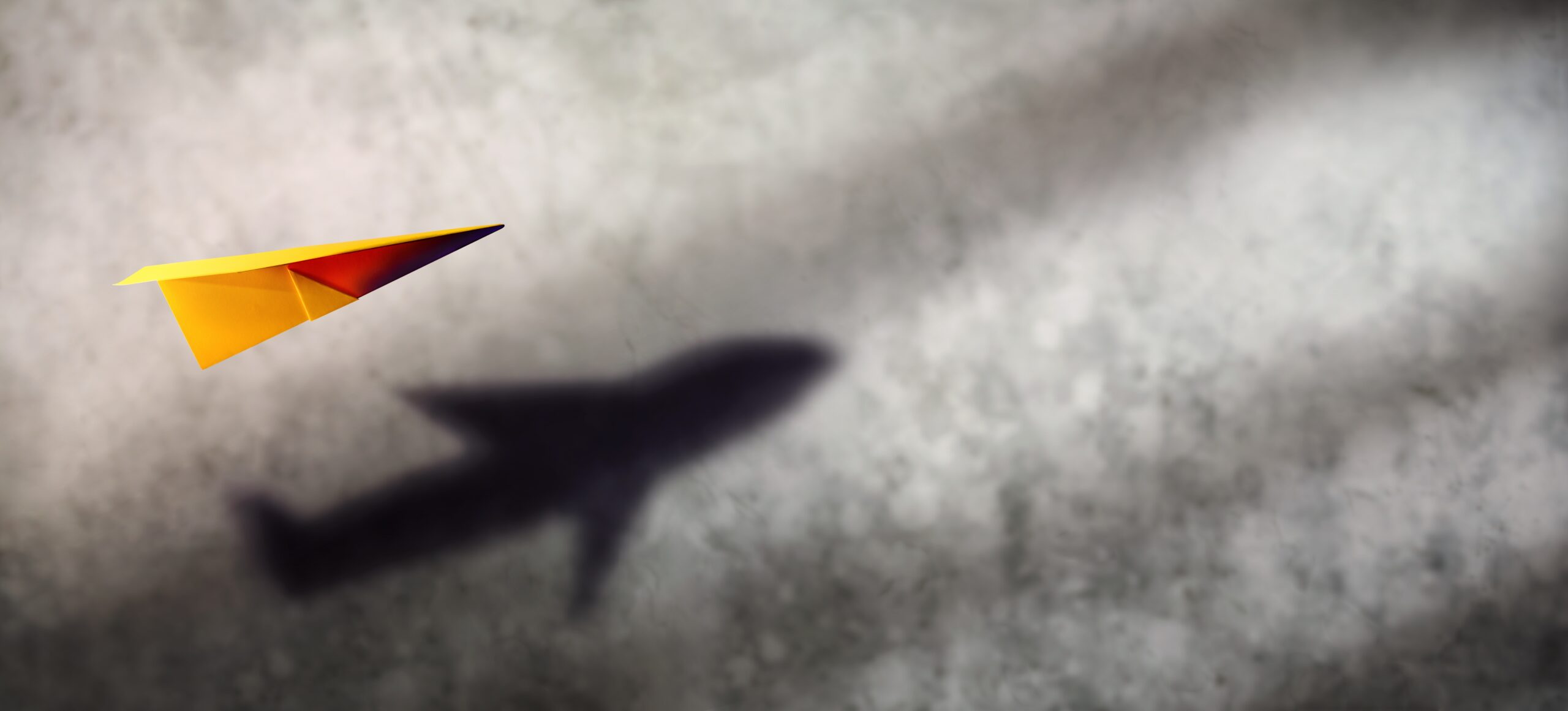 Paper Plane with Plane Shadow – A yellow paper plane casts the shadow of a real airplane on a cloudy wall.
