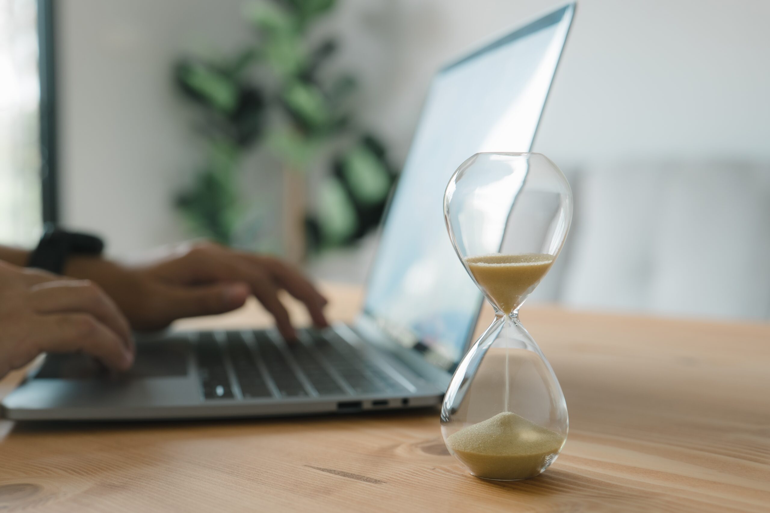 Close-up of an hourglass on a desk with a person working on a laptop in the background.