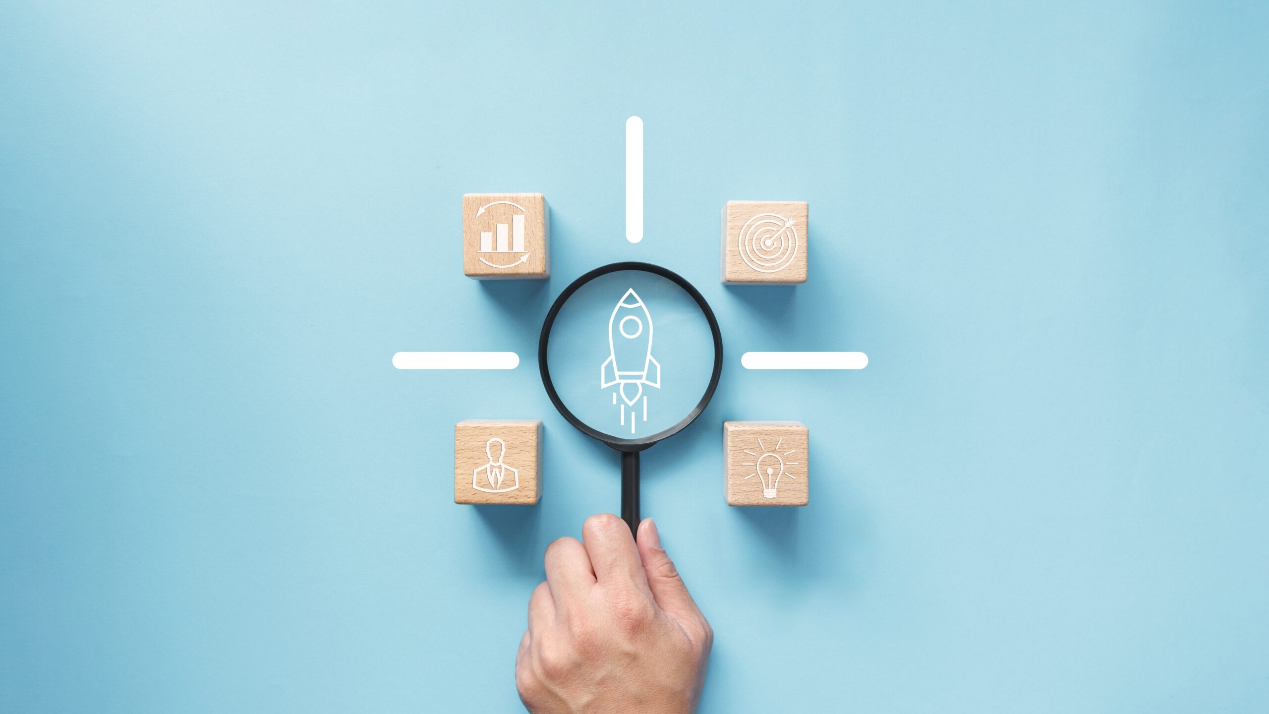 Hand holding magnifying glass over a startup icon surrounded by business symbols.